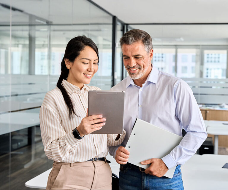 man and woman and office looking at tablet together. The man is holding some files